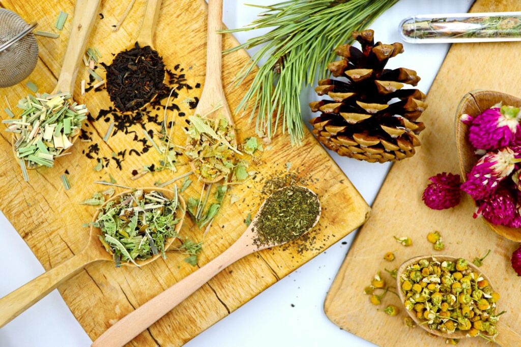 Lợi ích của trà thảo mộc - Hình ảnh nhìn từ trên xuống của nhiều loại trà thảo mộc pha trộn trên thìa gỗ với thông và hoa, trưng bày các thành phần tự nhiên.
