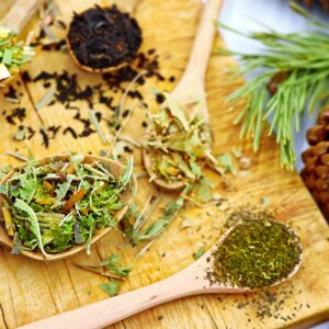 trà kết hợp siêu thực phẩm - Nhiều loại trà thảo mộc được trưng bày trên những chiếc thìa gỗ, đặt trên một tấm ván gỗ mộc mạc có quả thông.